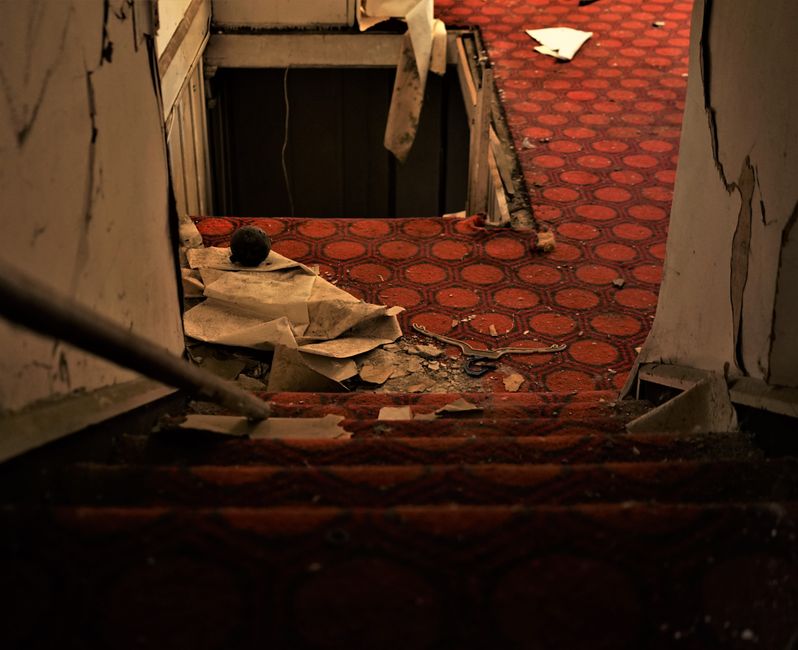 Inside a residential property during a home relocation, a staircase with red patterned carpet leads down to a lower level where a partly dismantled doorway reveals a dark hole or hatch, possibly for m