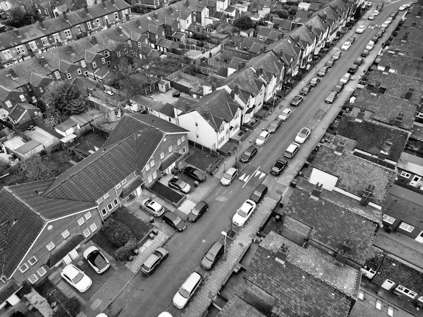 An aerial view in black and white showing a residential street lined with closely spaced houses, many with tiled or pitched roofs, and parked cars along both sides of the road. Some houses have small